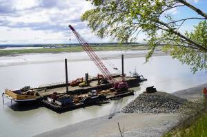 The barge, crane, and first pile of rock for the Kenai Bluff Stabilization Project is seen during a break in work at the bank of the Kenai River in Kenai, Alaska, on Tuesday, May 27, 2025. (Jake Dye/Peninsula Clarion)