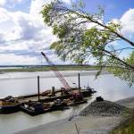 The barge, crane, and first pile of rock for the Kenai Bluff Stabilization Project is seen during a break in work at the bank of the Kenai River in Kenai, Alaska, on Tuesday, May 27, 2025. (Jake Dye/Peninsula Clarion)