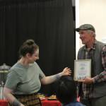 Homer Council on the Arts marketing coordinator Jenna Gerrety hands the Jill Berryman Arts Education Award to Bill Worsfold, who accepted it on behalf of recipient Shellie Worsfold, at Homer Council on the Arts annual Community Arts Award Ceremony on Saturday, July 19, 2025, in Homer, Alaska. (Delcenia Cosman/Homer News)