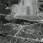 This 1960 aerial photograph from the Civil Aeronautics Administration shows, from top to bottom, the southern end of the Kenai Airport runway, the Spur Highway and the fledgling city of Kenai. Near the center of the photo is a cluster of white-painted buildings then owned by the U.S. Fish & Wildlife Service, which managed the Kenai National Moose Range. Enclosed in a small group of trees south-southwest of these buildings is the Russian Orthodox Church.