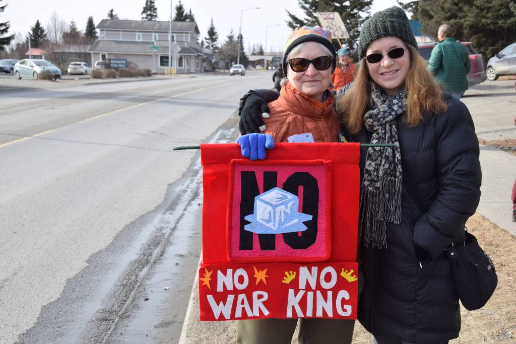 Kippy Dalton (right) and an anonymous community member pose on the sidewalk along Pioneer Avenue with a handmade quilted banner that reads, No (ICE), No War, No King, during the No Kings protest on Saturday, March 28, 2026, in Homer, Alaska. (Delcenia Cosman/Homer News)
